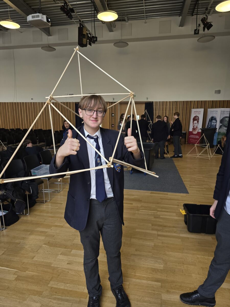 A student in a blazer and glasses smiles and gives two thumbs up while standing behind a medium-sized wooden tetrahedron frame he has helped build.