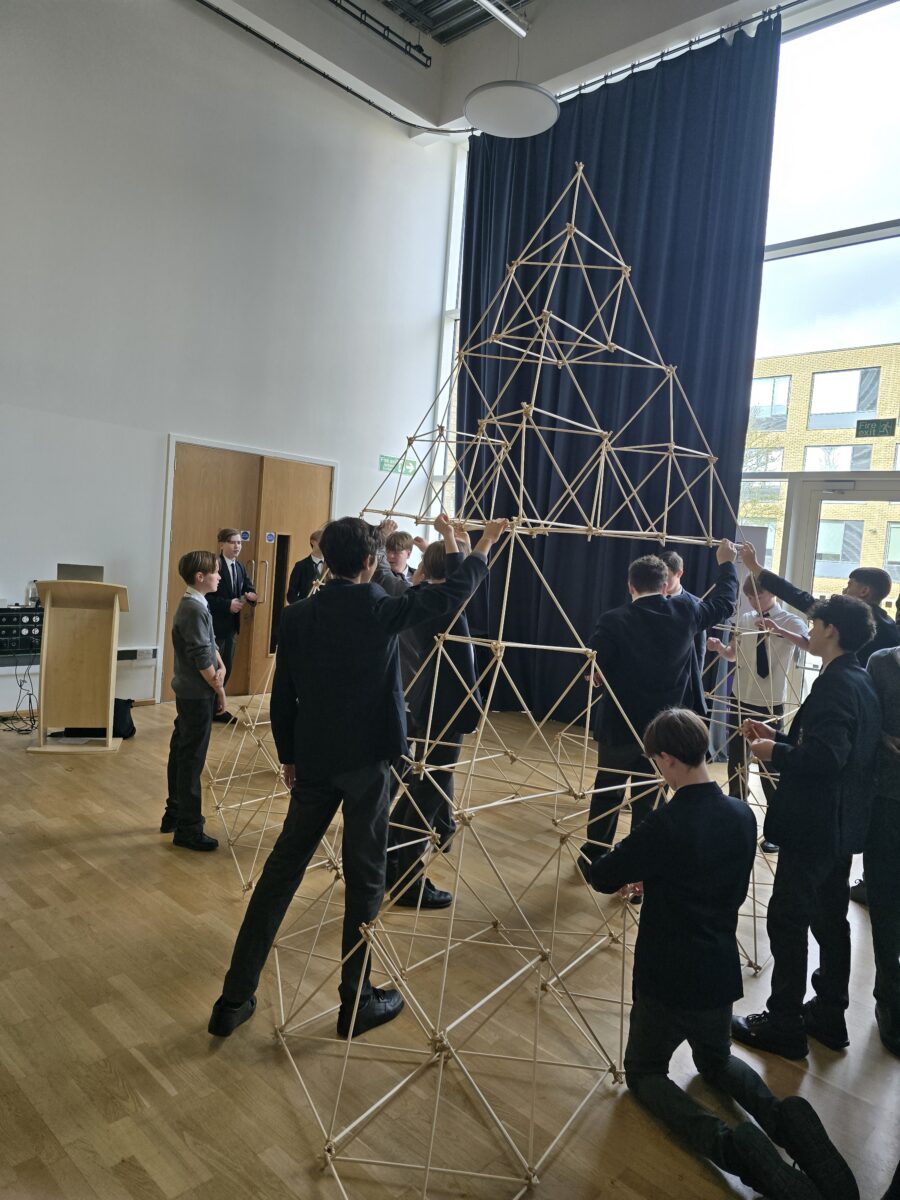 A large group of students stands around a massive, multi-tiered pyramid structure made of hundreds of wooden dowels, reaching toward the ceiling of the hall as they add the final sections.