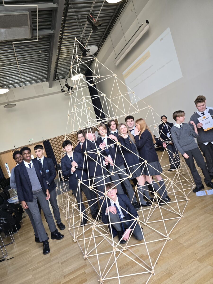 A wide shot of a diverse group of students posing proudly behind and inside the hollow sections of a giant, completed wooden tetrahedron structure that stands taller than them.
