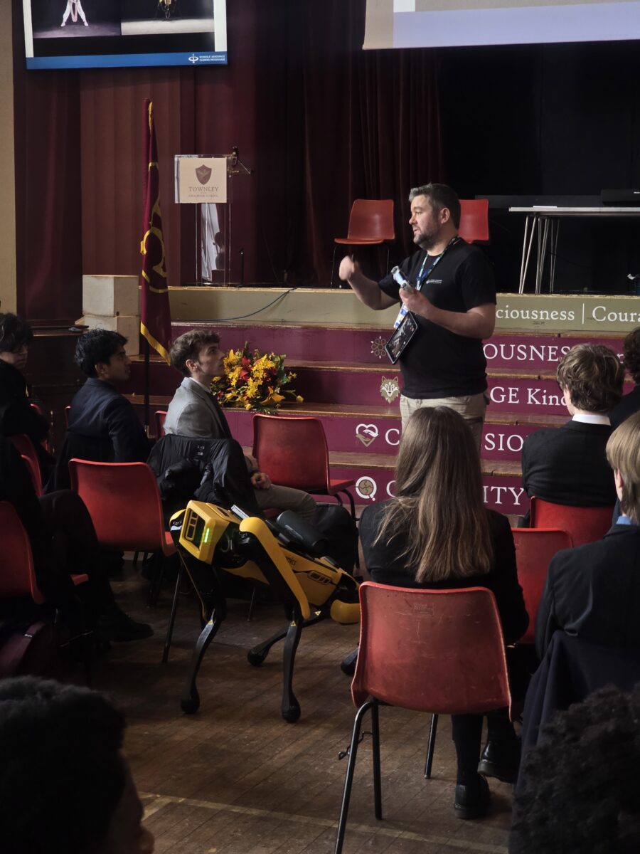 A man standing on a stage speaking and holding a microphone, with a yellow robot dog standing on the floor in front of an audience of seated students.