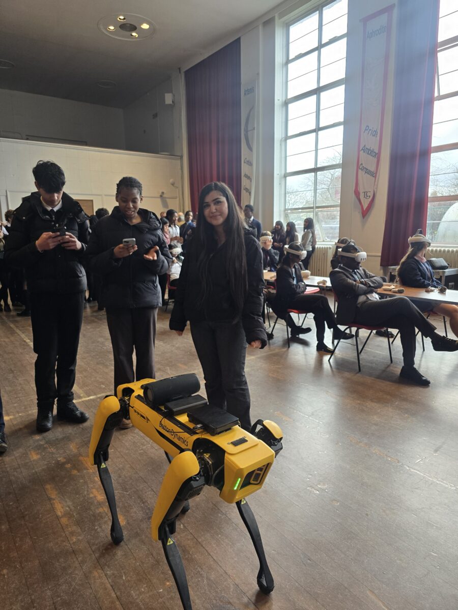 A yellow Boston Dynamics robot dog standing on a wooden floor, with three students standing behind it looking at their phones, and other students seated in the background wearing VR headsets.