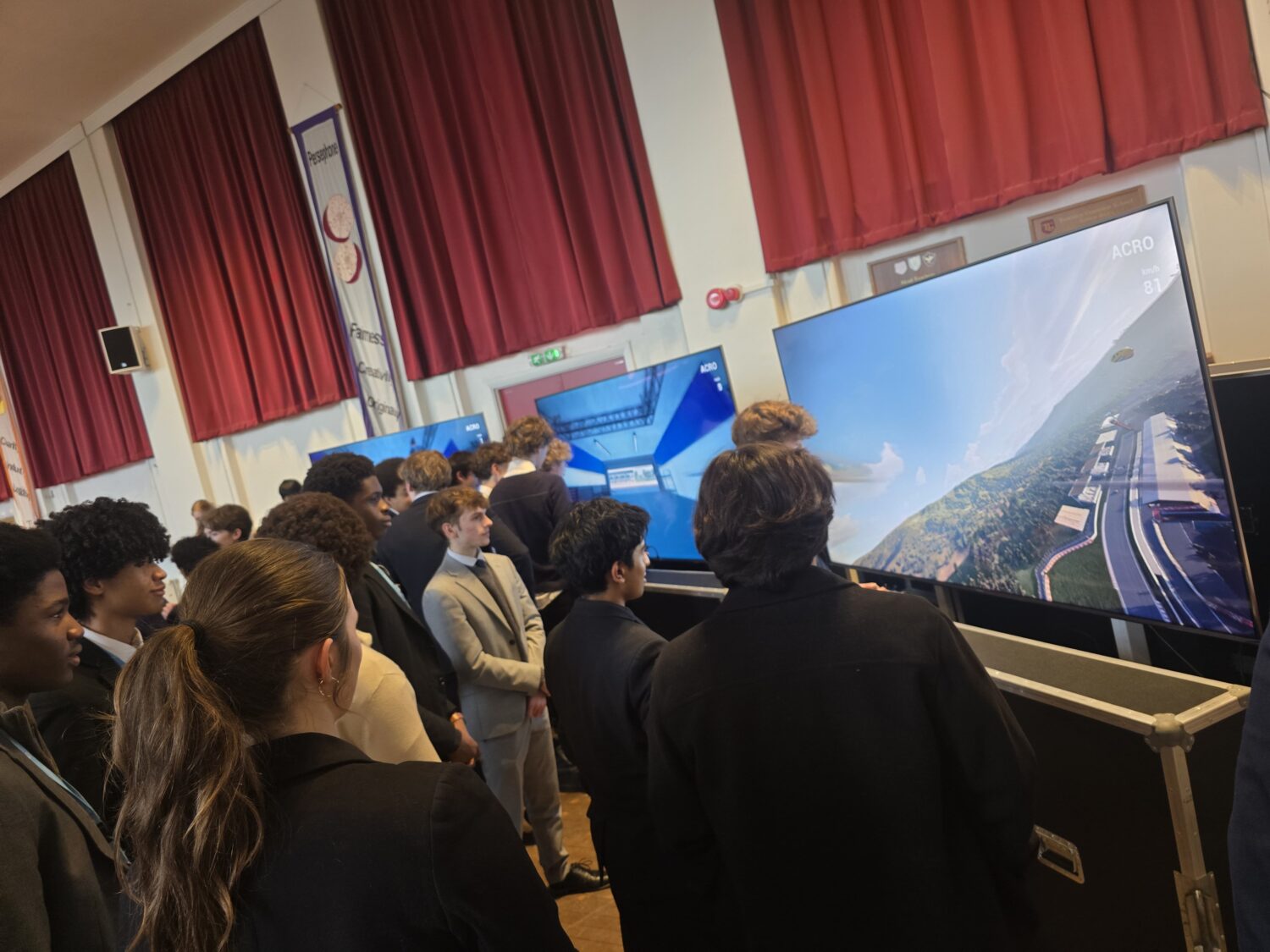 A group of students observing large screens displaying a flight simulator program, showing an aerial view of a landscape with a runway.