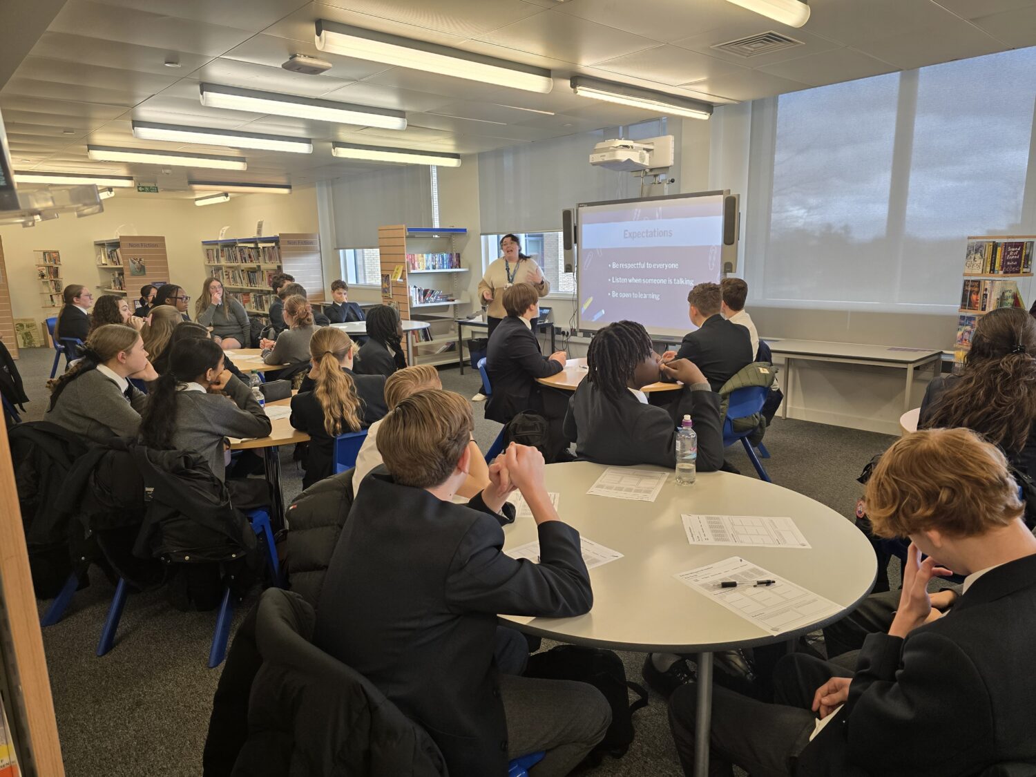 A wide view of a library setting where a woman presents to a group of students seated at tables with papers and pens in front of them.