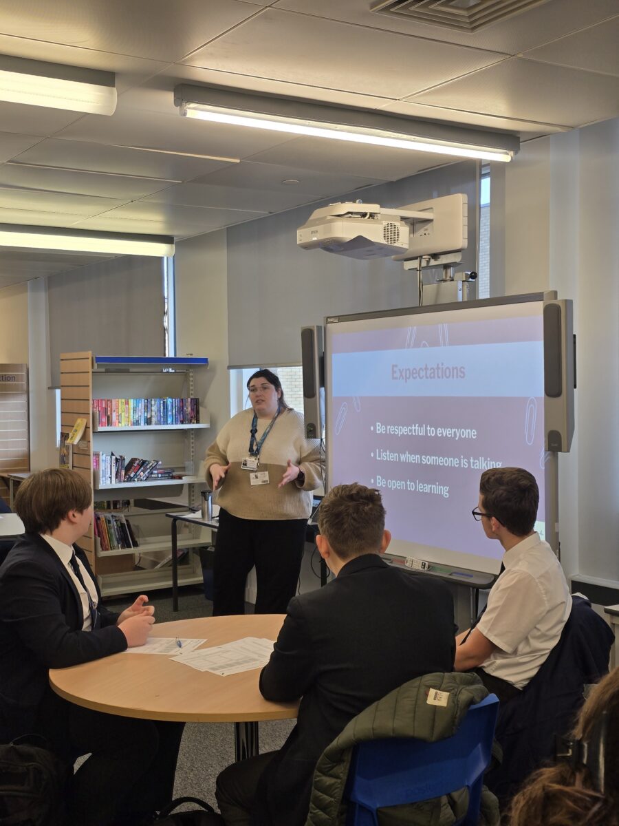 A presenter stands next to a screen displaying "Expectations" while speaking to three students seated at a round table in a library.
