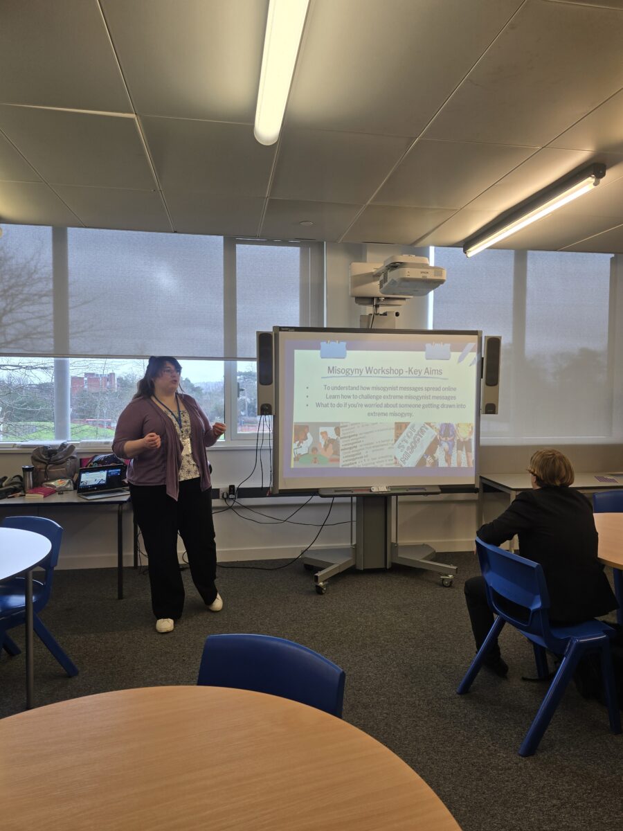 A woman gestures with her hands while standing in front of a presentation screen displaying a slide titled "Misogyny Workshop - Key Aims."