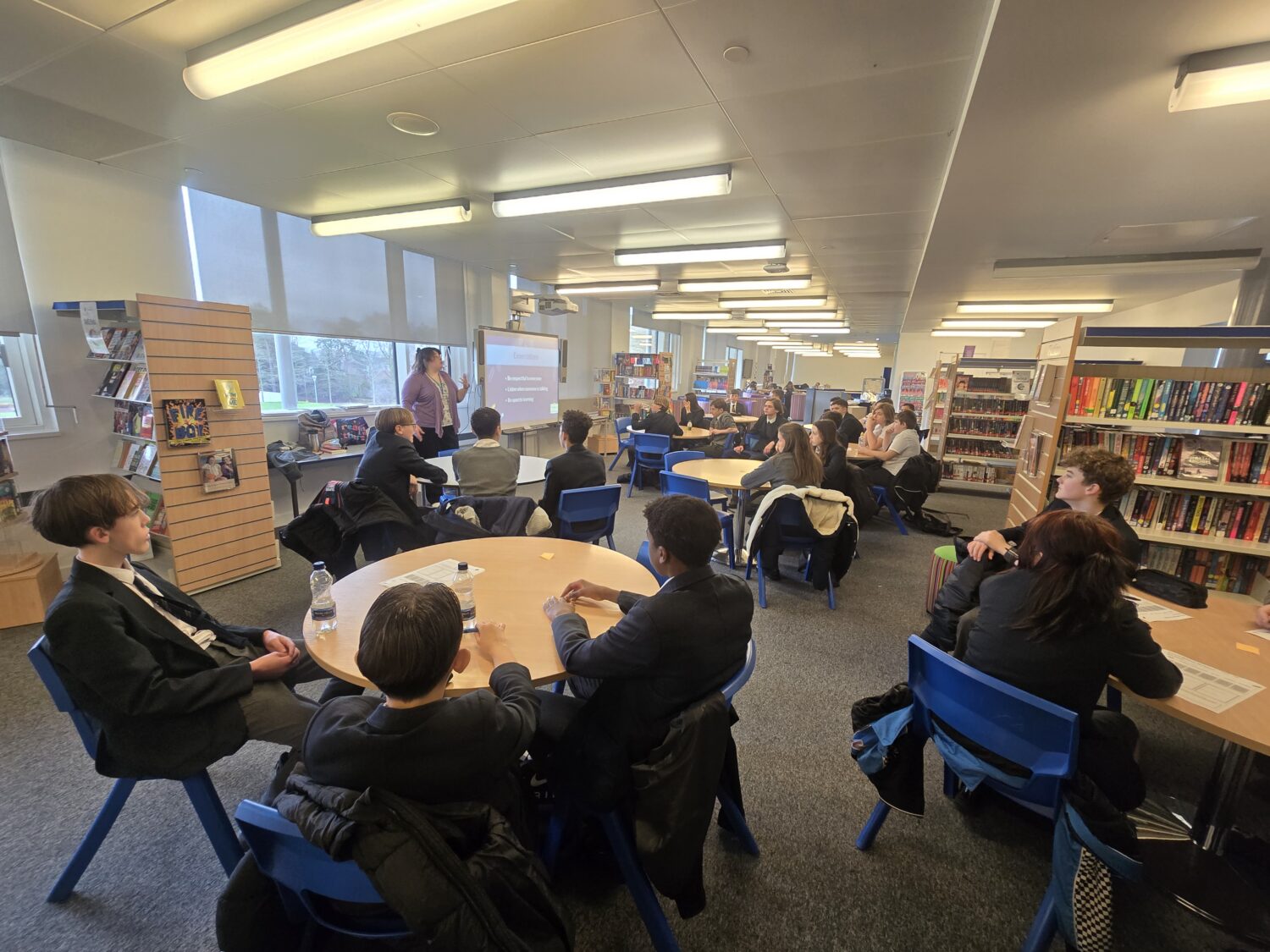 Students in school uniforms sit at round tables in a library, listening to a presentation given by a woman standing near a digital screen.