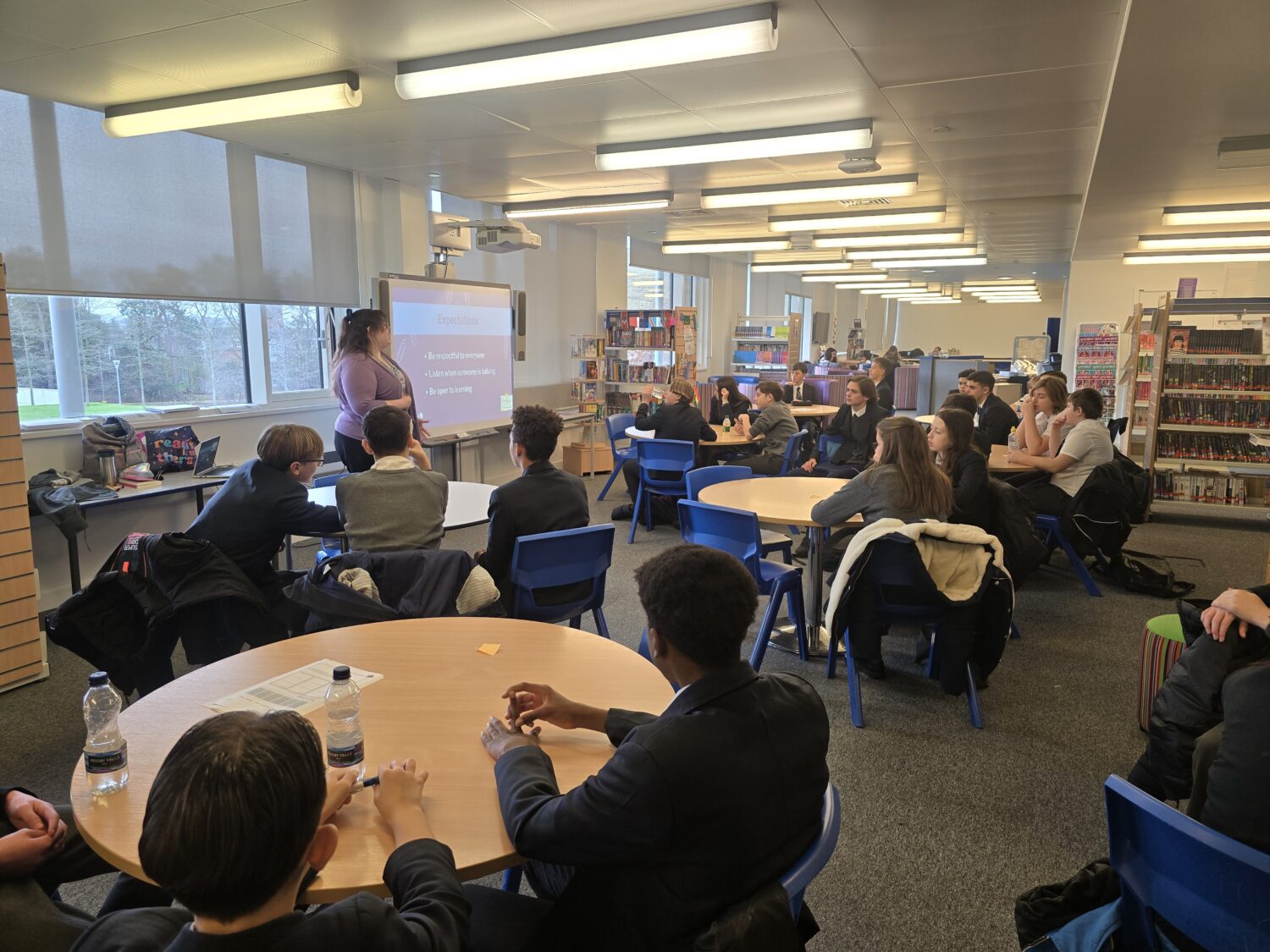 A woman stands by a screen titled "Expectations" presenting to a room of uniformed students seated at round tables in a library.