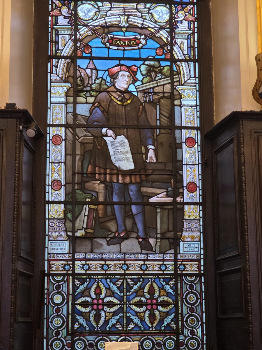 A vertical stained-glass window depicting a man in historical attire labeled "CAXTON" holding a sheet of paper, surrounded by ornate decorative borders and floral patterns.