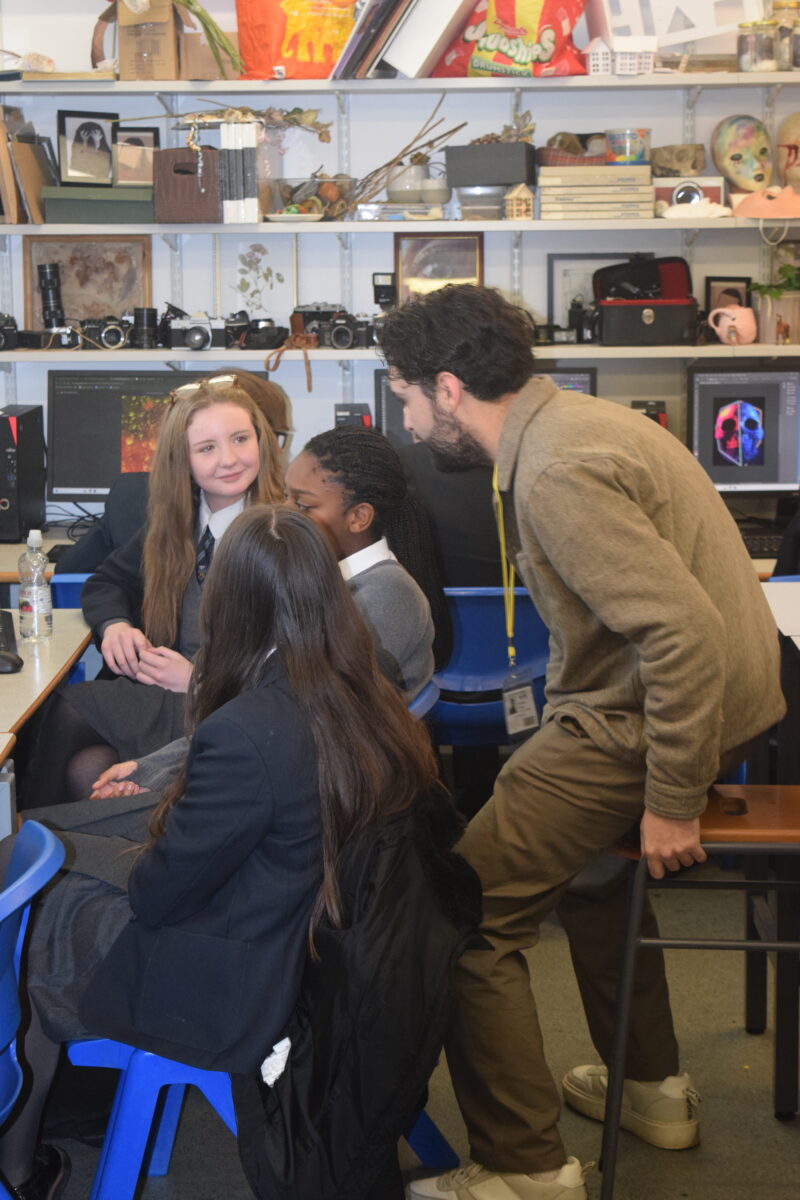 A teacher leans in to engage in conversation with three female students seated together in an art classroom filled with photography equipment and supplies.