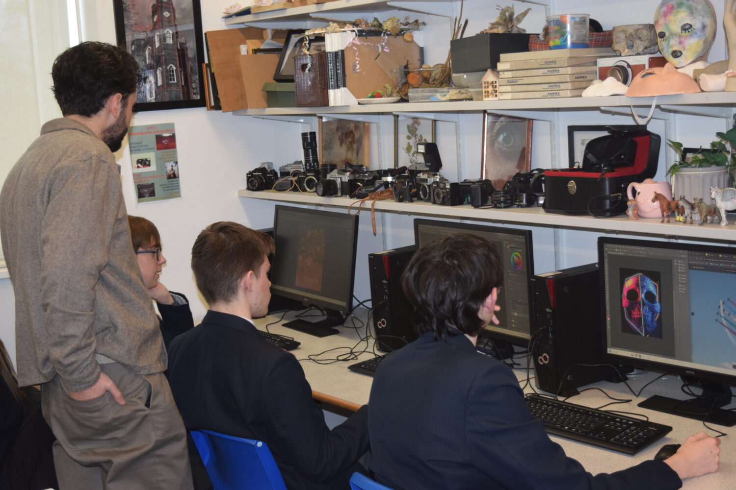 A teacher stands behind three uniformed students working on digital art projects on computers in a classroom, with shelves above lined with vintage cameras and eclectic art props.