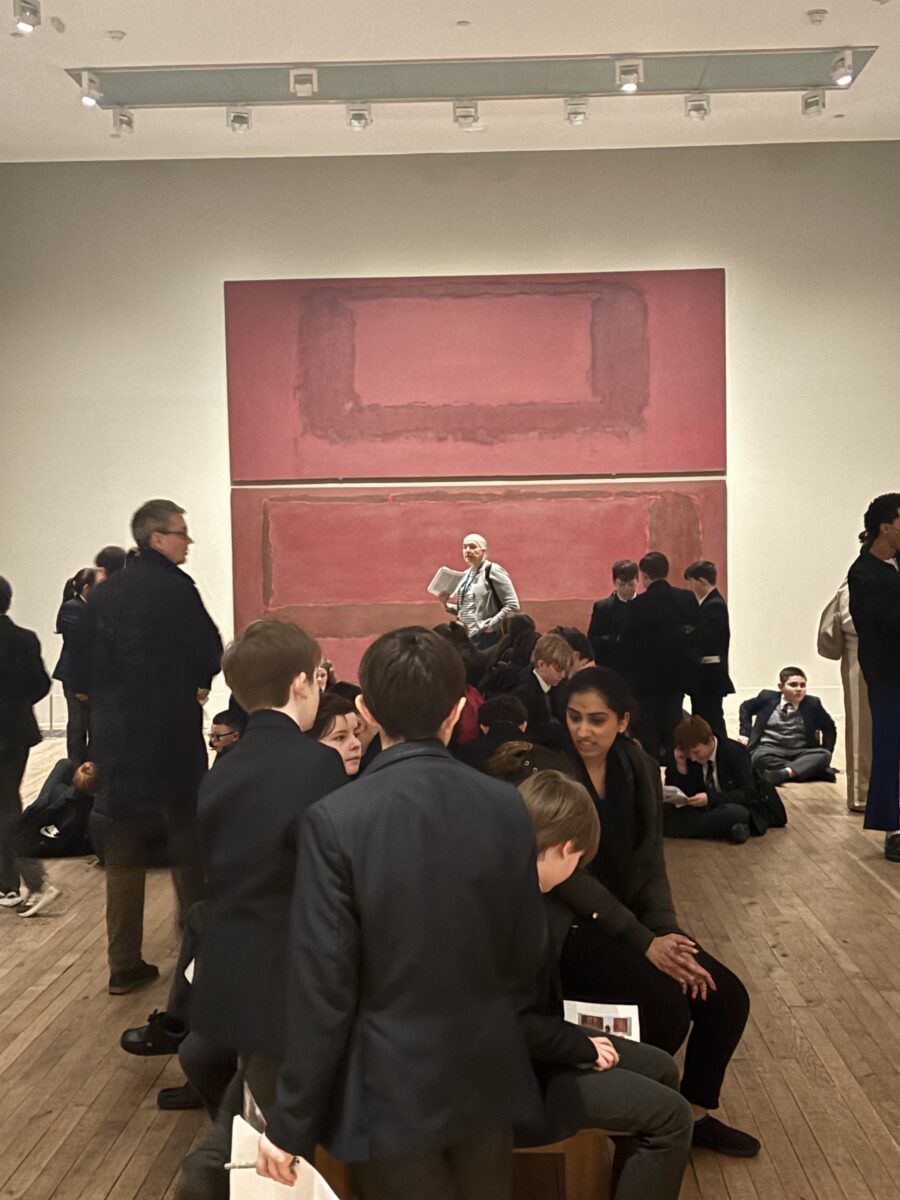 A group of students and adults inside an art gallery with wooden floors, standing and sitting in front of large, red-toned abstract color field paintings.