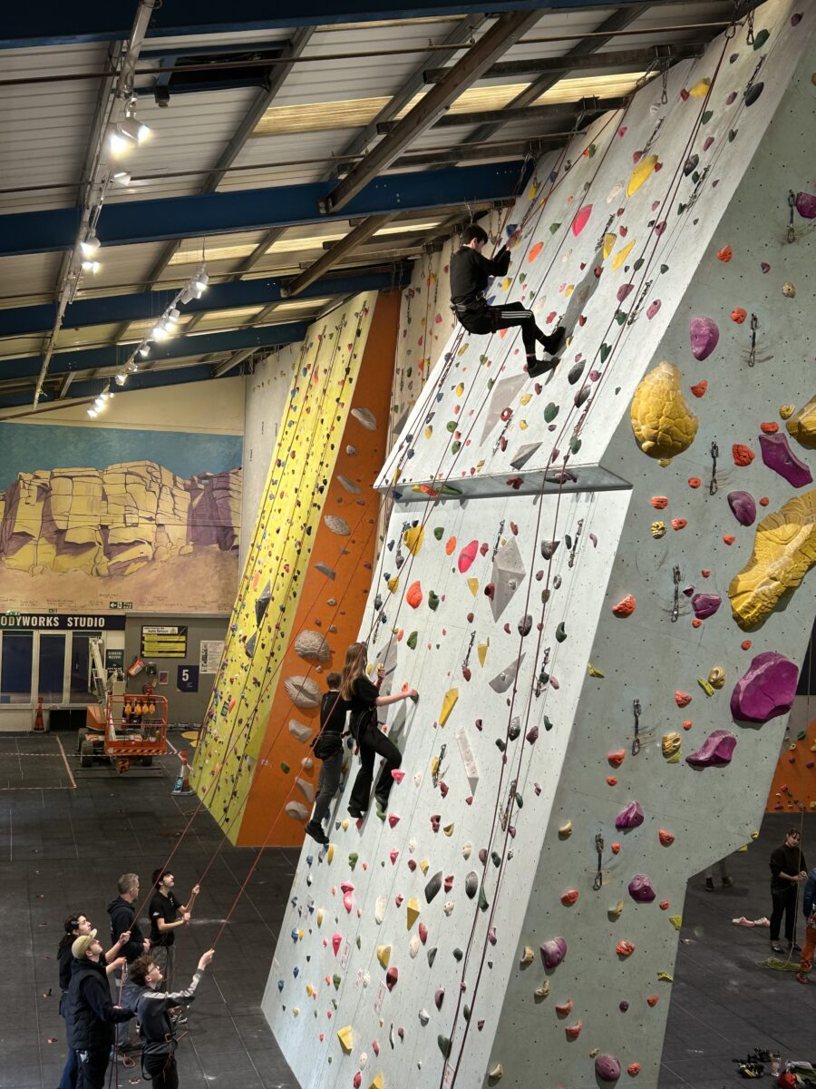 Three people ascend an indoor climbing wall while five people look on