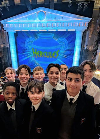 A group of smiling students in school uniforms posing in a theatre balcony, with a bright blue stage set behind them featuring a Greek temple design and a large vase labeled "HERCULES."