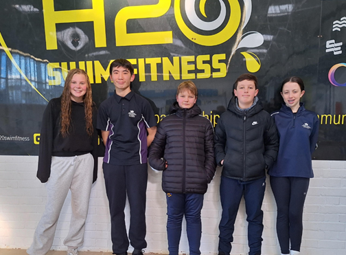 Five smiling students standing in a row wearing athletic clothing and school sports kit in front of a large banner that reads "H2O SWIM FITNESS."