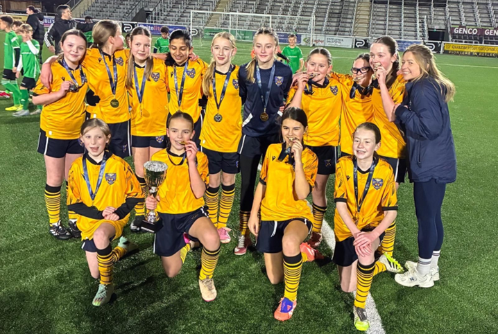 On a football pitch, a girls football team, in a yellow kit picture with medals and a trophy celebrate with two coaches.