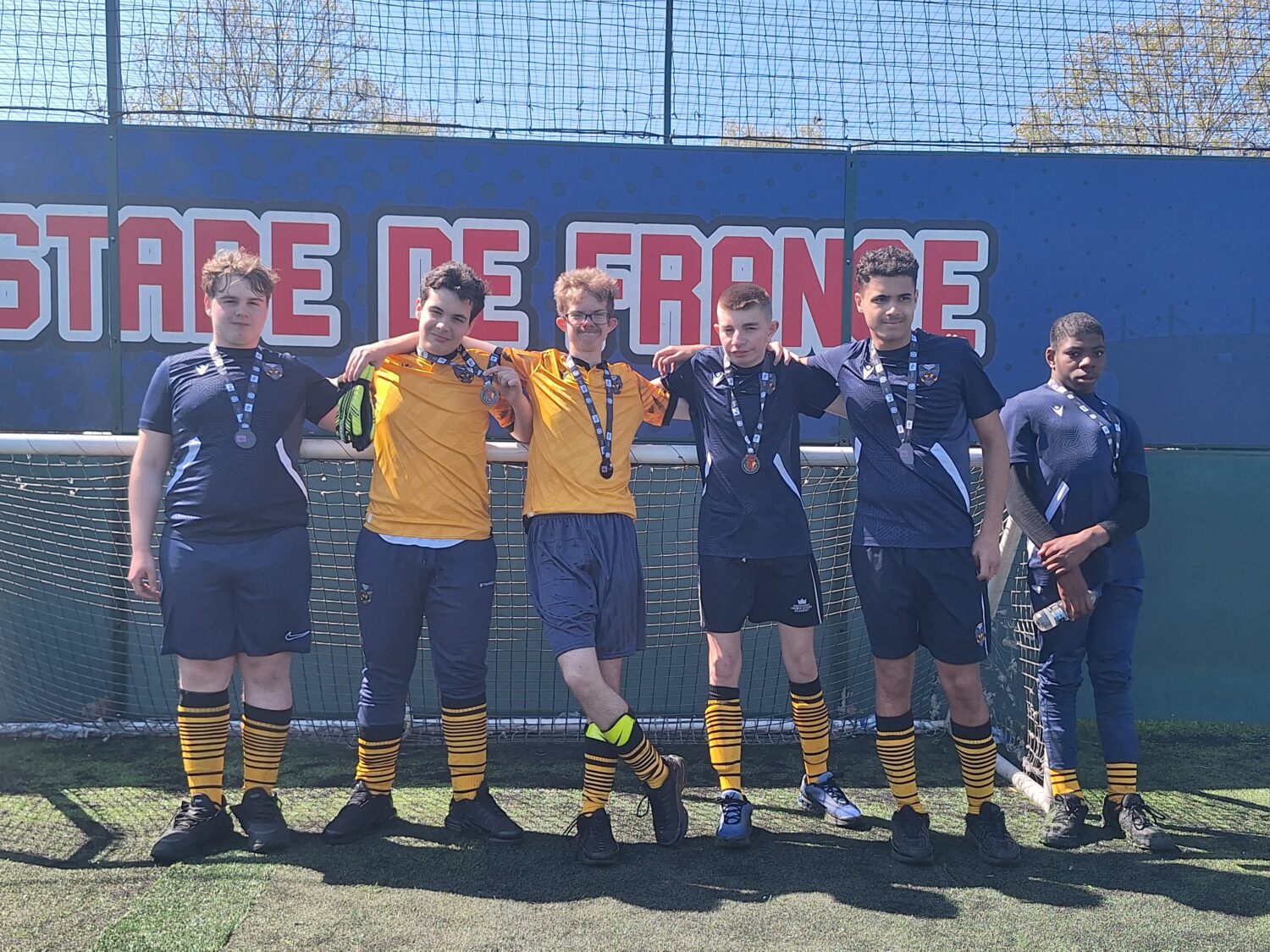 Six people in soccer uniforms standing shoulder-to-shoulder with their arms around each other in front of a "Stade de France" banner.