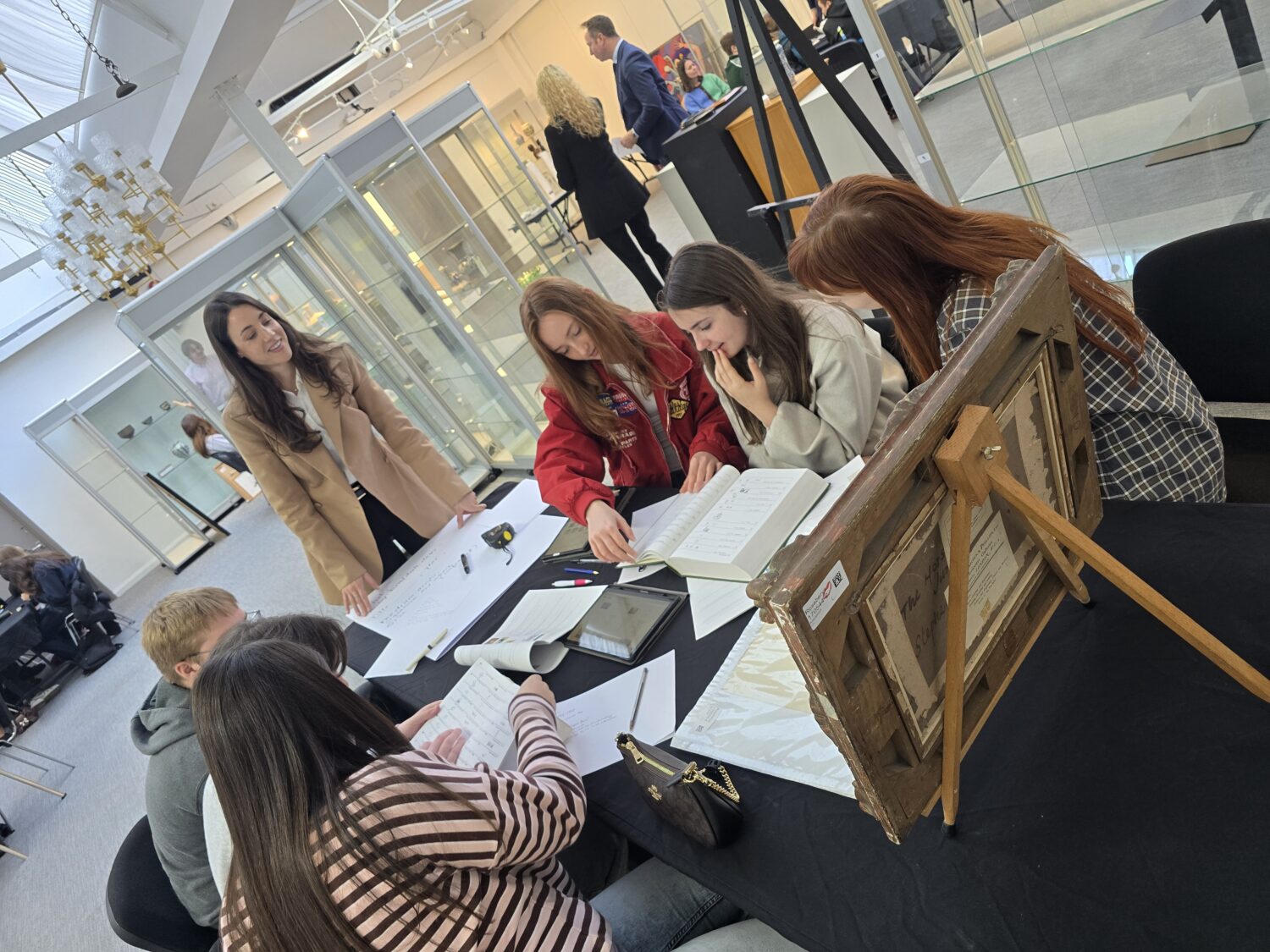 A group gathered around a black table examining paperwork and the back of a framed artwork resting on a small wooden easel.