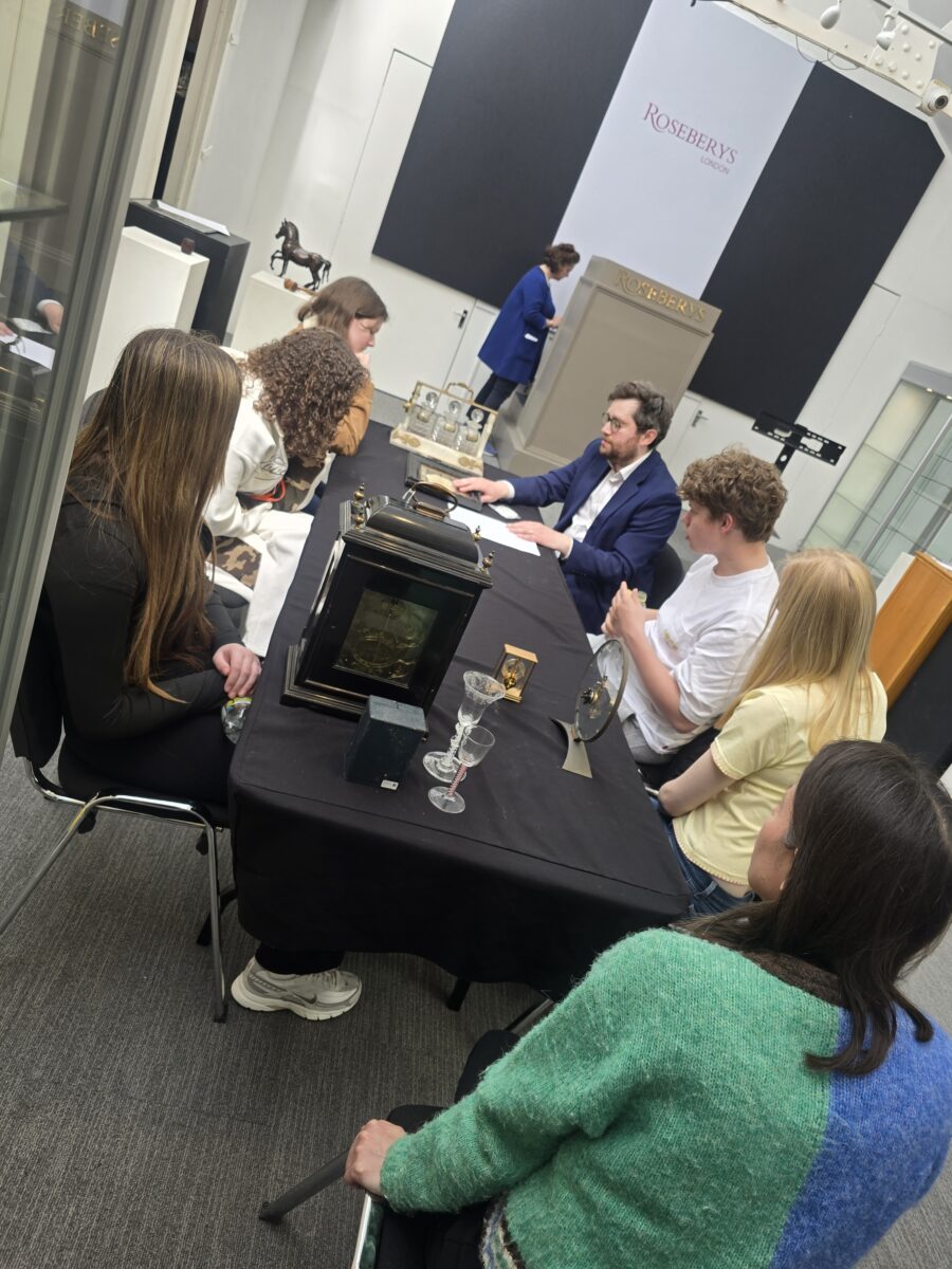 Several people seated around a table looking at antique objects, including an old clock and glassware, while a man in a blue suit speaks with them.