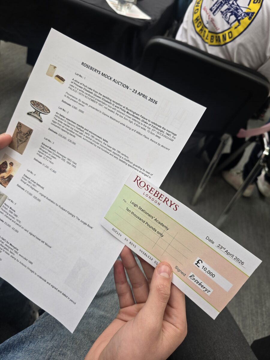 A close-up of a person holding a printed sheet for a "Roseberys Mock Auction" alongside a novelty cheque for £10,000 made out to Leigh Stationers' Academy.