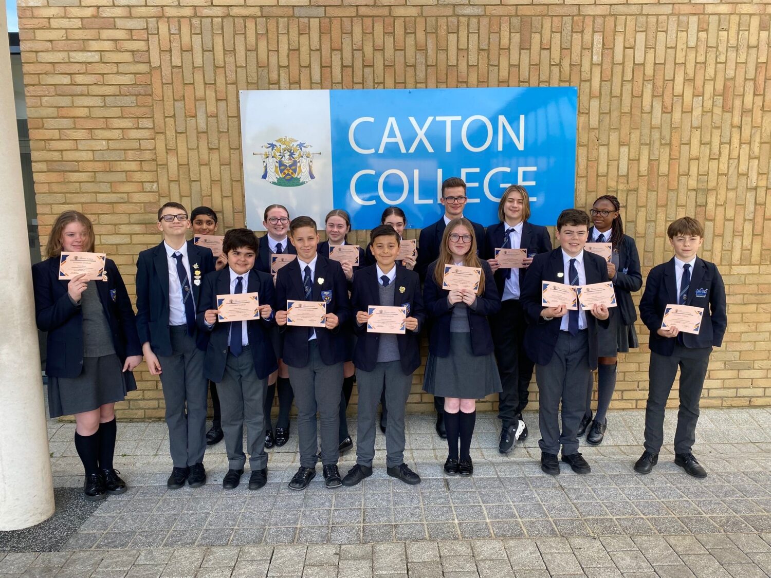 A group of people in school uniforms posing outdoors with certificates in front of a bright blue sign that reads "Caxton College."