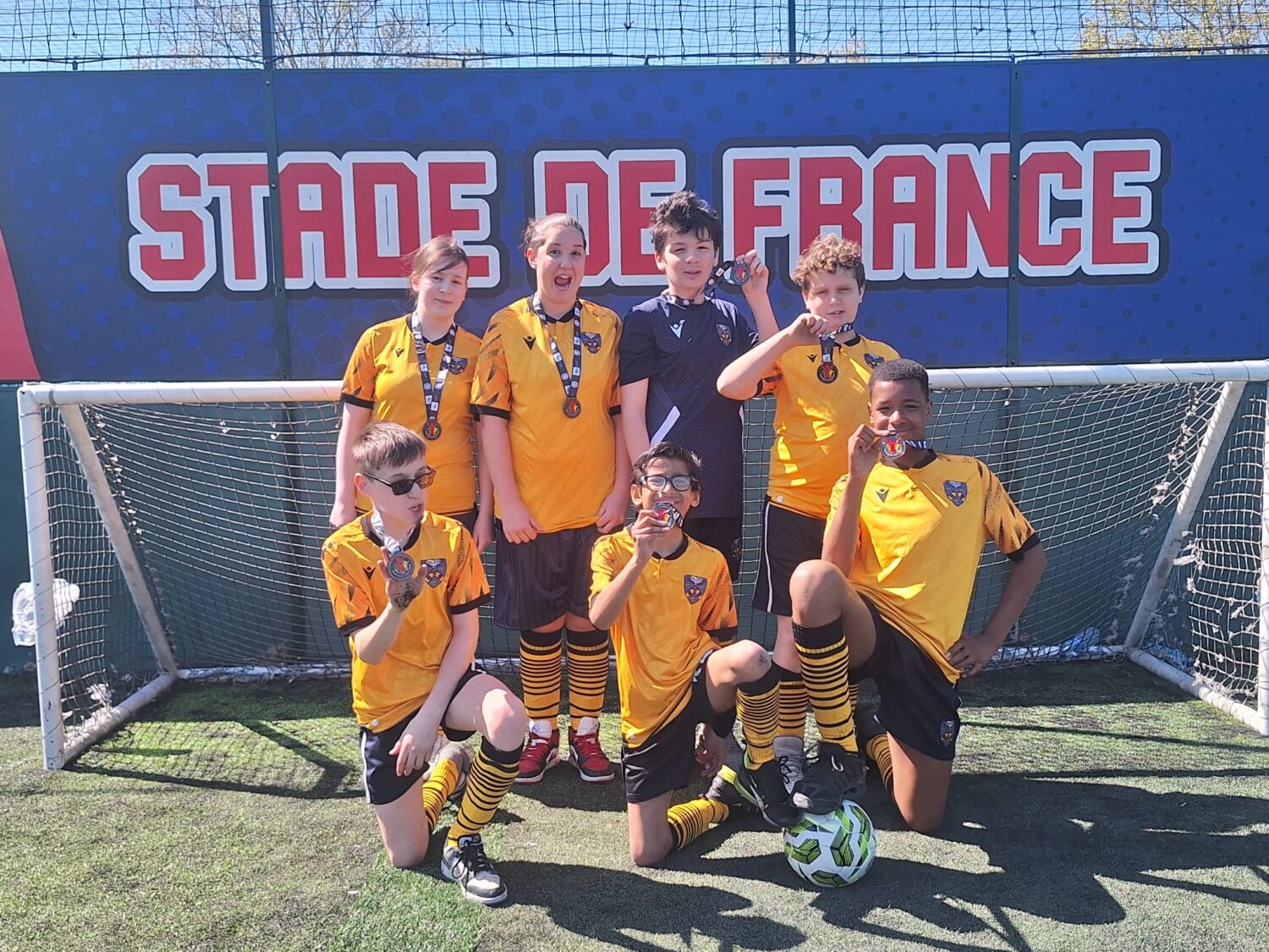 A group of people in yellow and black soccer uniforms posing on a turf field with medals in front of a "Stade de France" banner