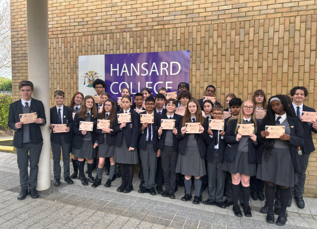 A group of people in school uniforms posing outdoors with certificates in front of a purple sign that reads "Hansard College."