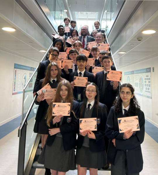 A group of people in school blazers and ties standing together on a modern indoor staircase, holding up printed certificates.