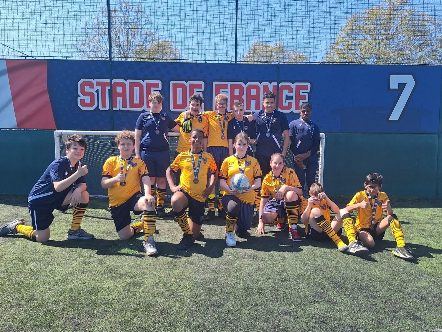 A large group of people in yellow, black, and navy soccer uniforms posing with medals and a soccer ball in front of a "Stade de France" banner.