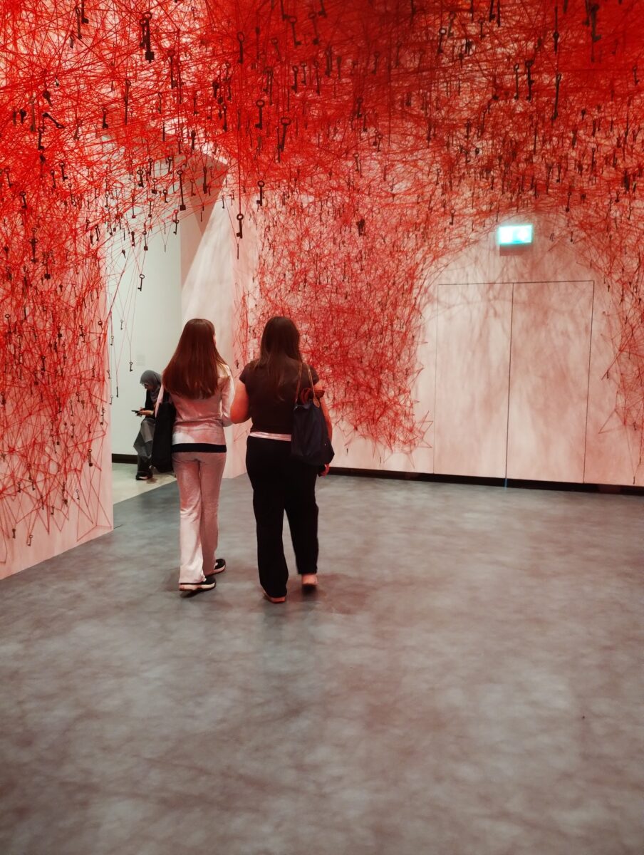 Two visitors walk toward an arched doorway formed by thousands of tangled red threads suspended from the ceiling. Hanging within the web are numerous vintage metal keys, creating a dense, cloud-like canopy.
