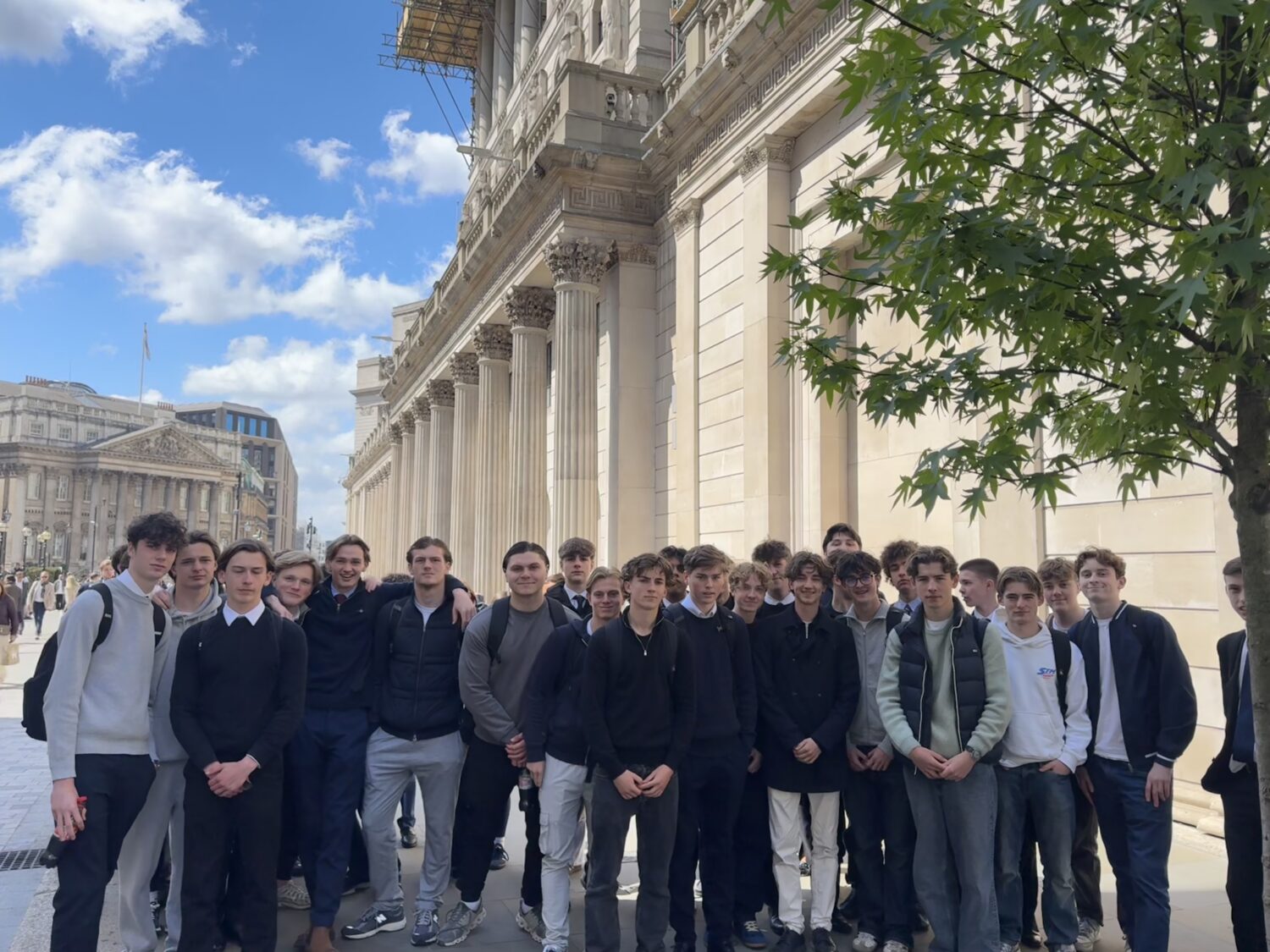 A large group of male students poses together in front of the grand, pillared facade of the Bank of England.