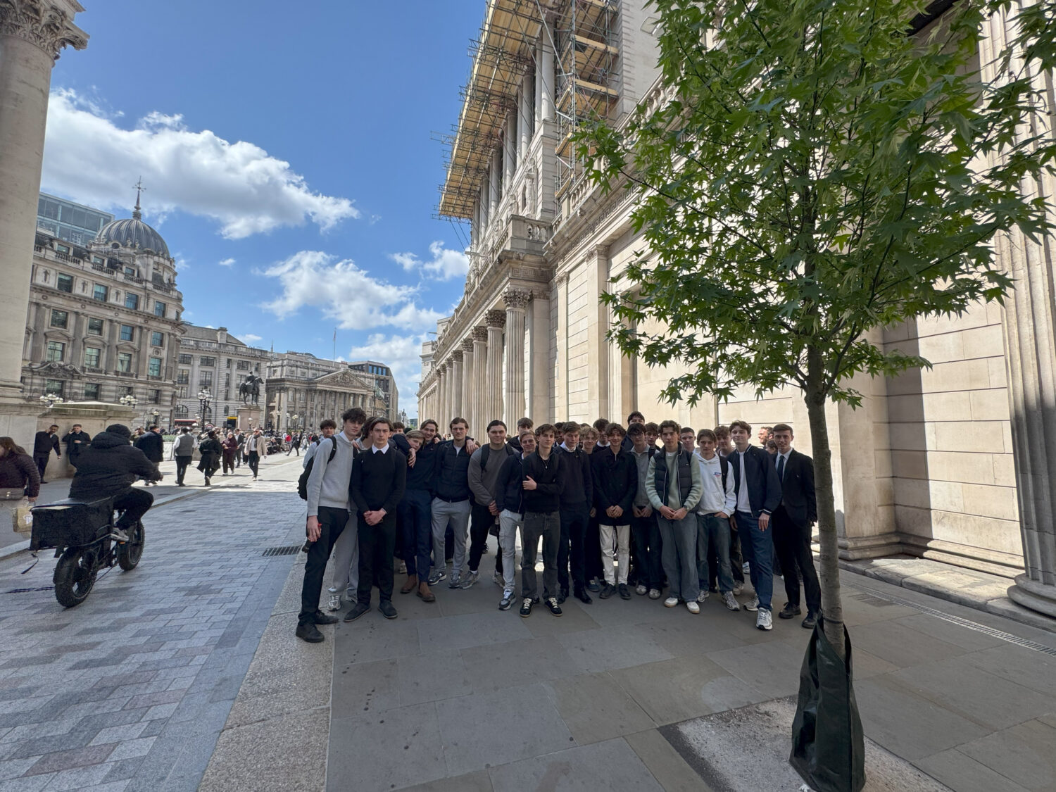 A large group of male students poses together in front of the grand, pillared facade of the Bank of England.
