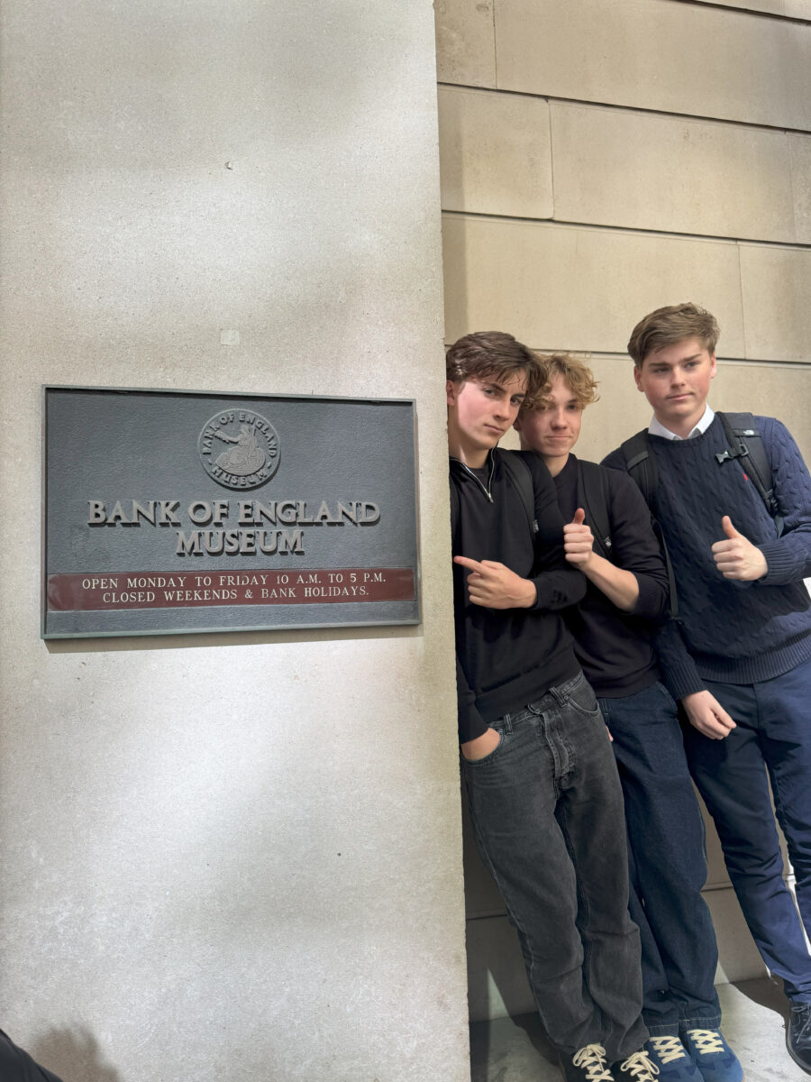 Three students stand next to a bronze "Bank of England Museum" sign; two give a thumbs-up and one points to the sign.