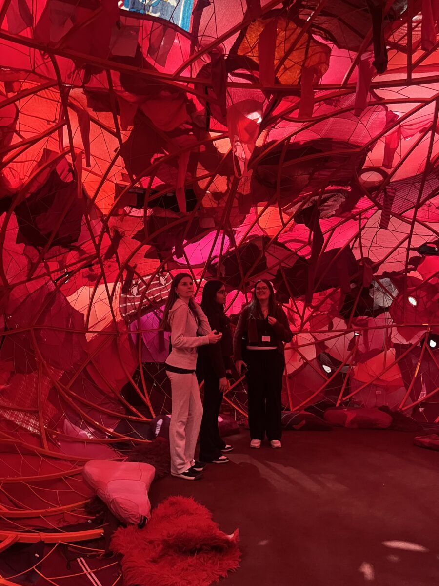 Three students stand inside a large, spherical structure made of a reddish-orange metal framework. The interior is layered with translucent fabrics and materials in shades of red and pink, creating a warm, glowing, cocoon-like atmosphere.