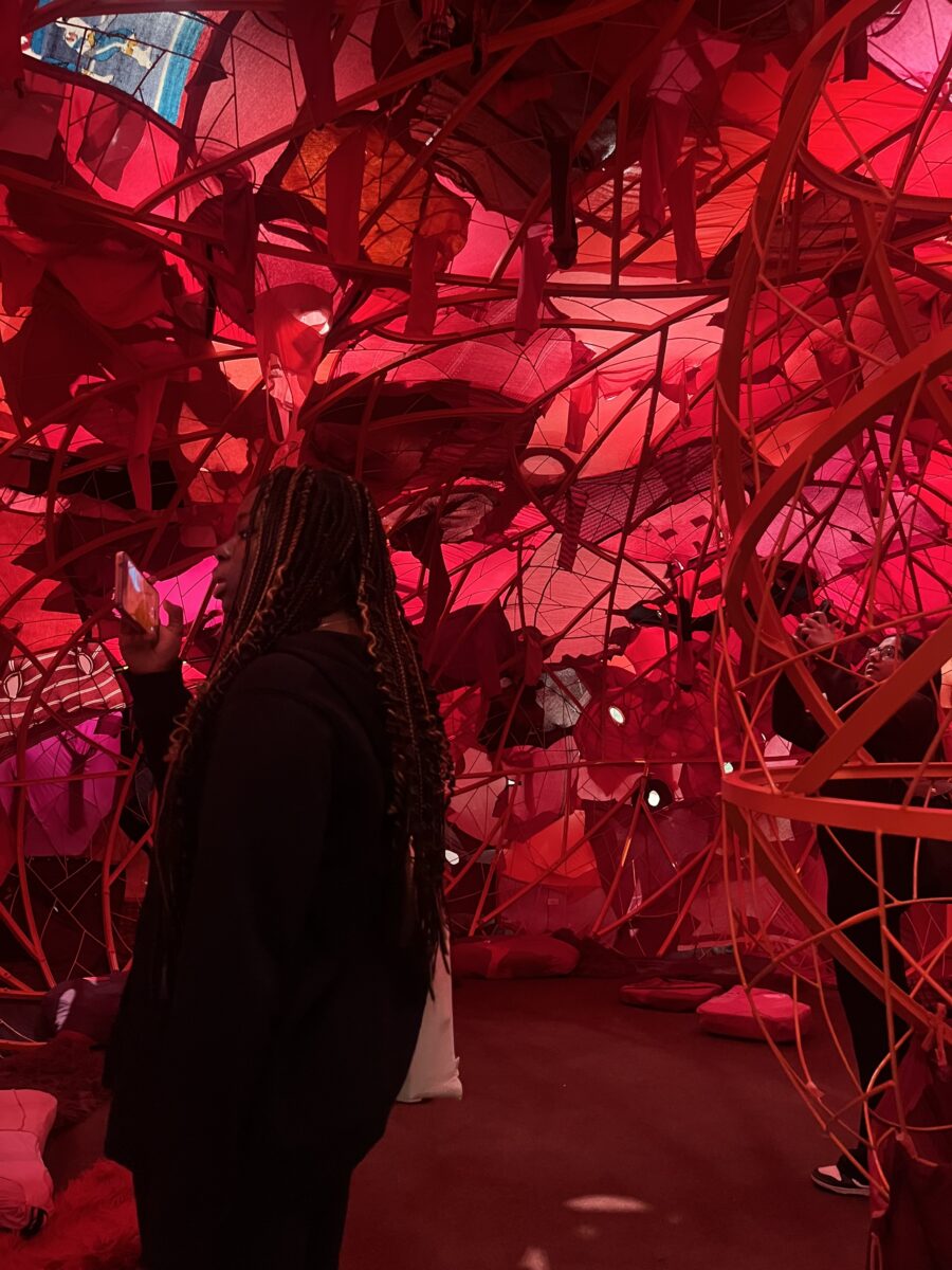 : A close-up view from inside the same spherical installation. A visitor with long braids is seen in profile, using her phone to take a photo of the complex geometric patterns and light filtering through the red layers.