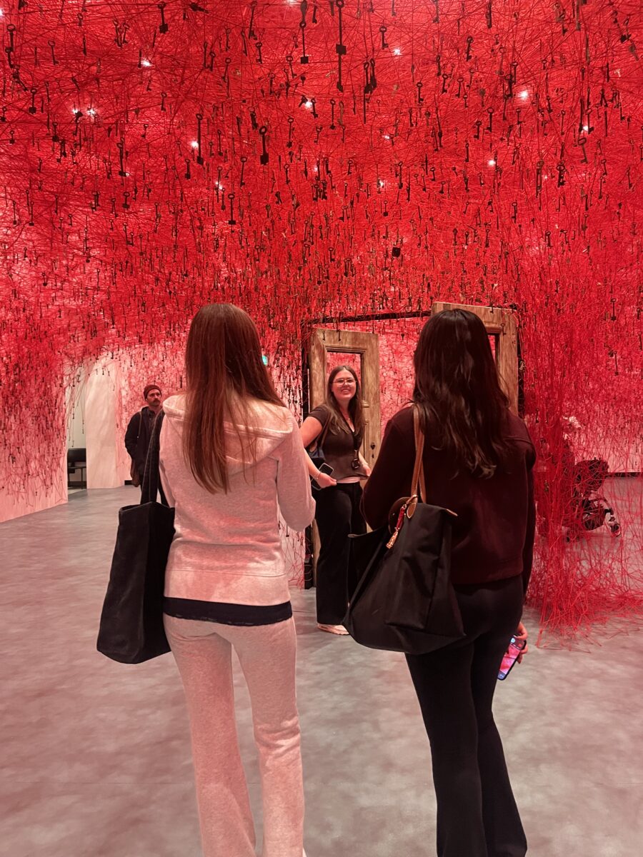 A wider view of the "Key" installation. Two students in the foreground look toward a third student standing in a free-standing wooden door frame. The entire room is saturated with the red thread network and suspended keys, symbolizing memory and interconnectedness.