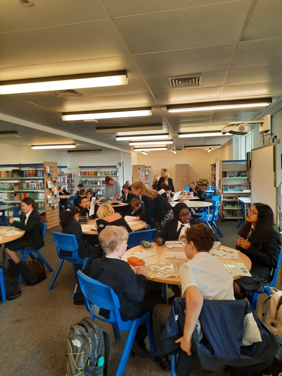 Students working in groups at round tables in the academy library, with bookshelves along the walls.