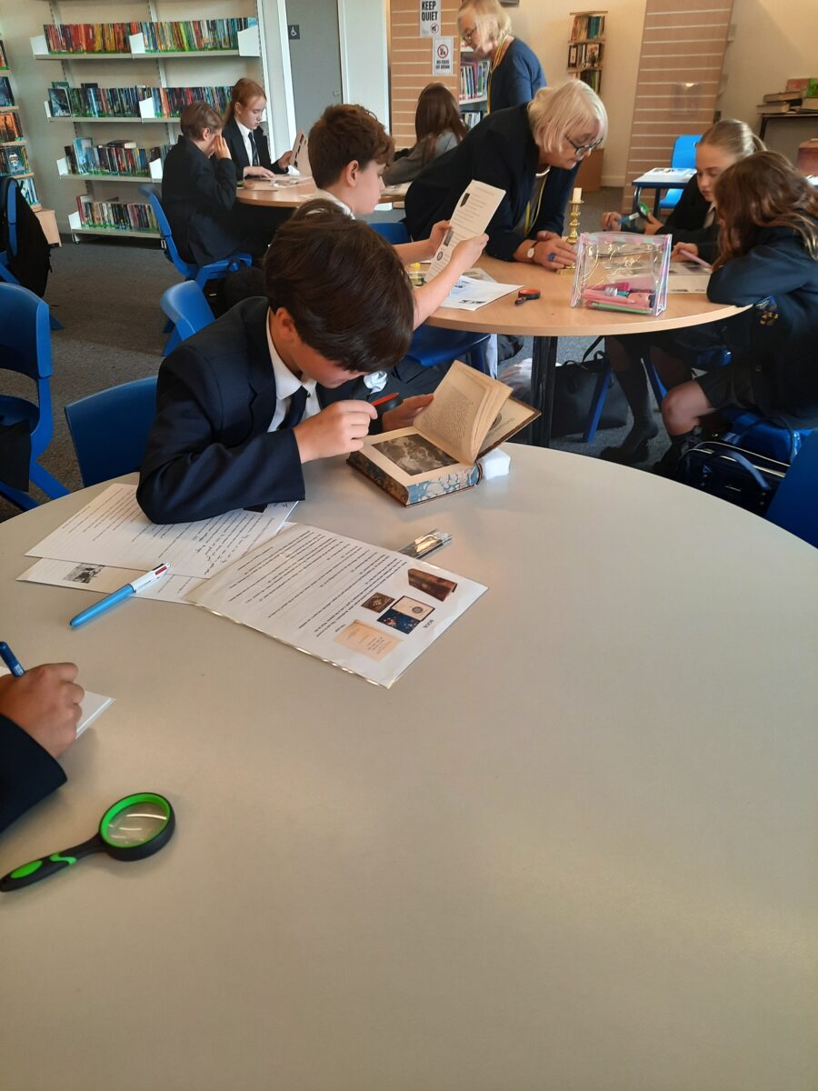 A student in school uniform leans over a table to examine historical items, while other students work nearby.