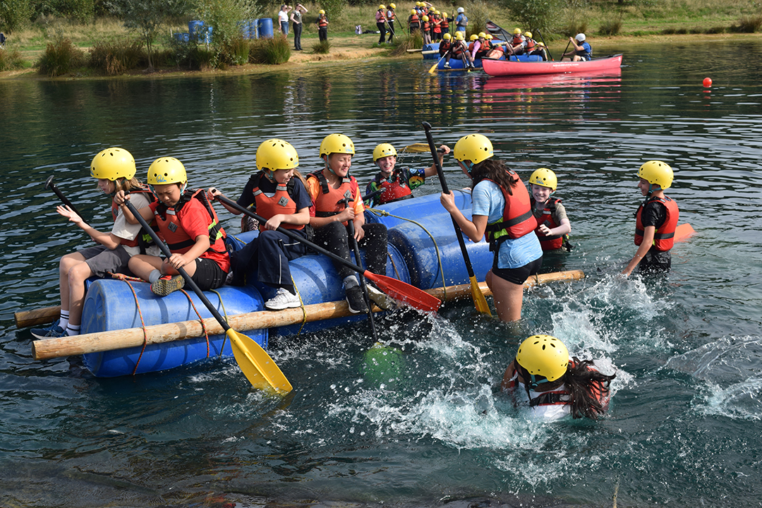 Nine children, wearing helmets and lifejackets, on a handmade raft made out of wood and plastic tubs. They are smiling and five have fallen off and are in the water