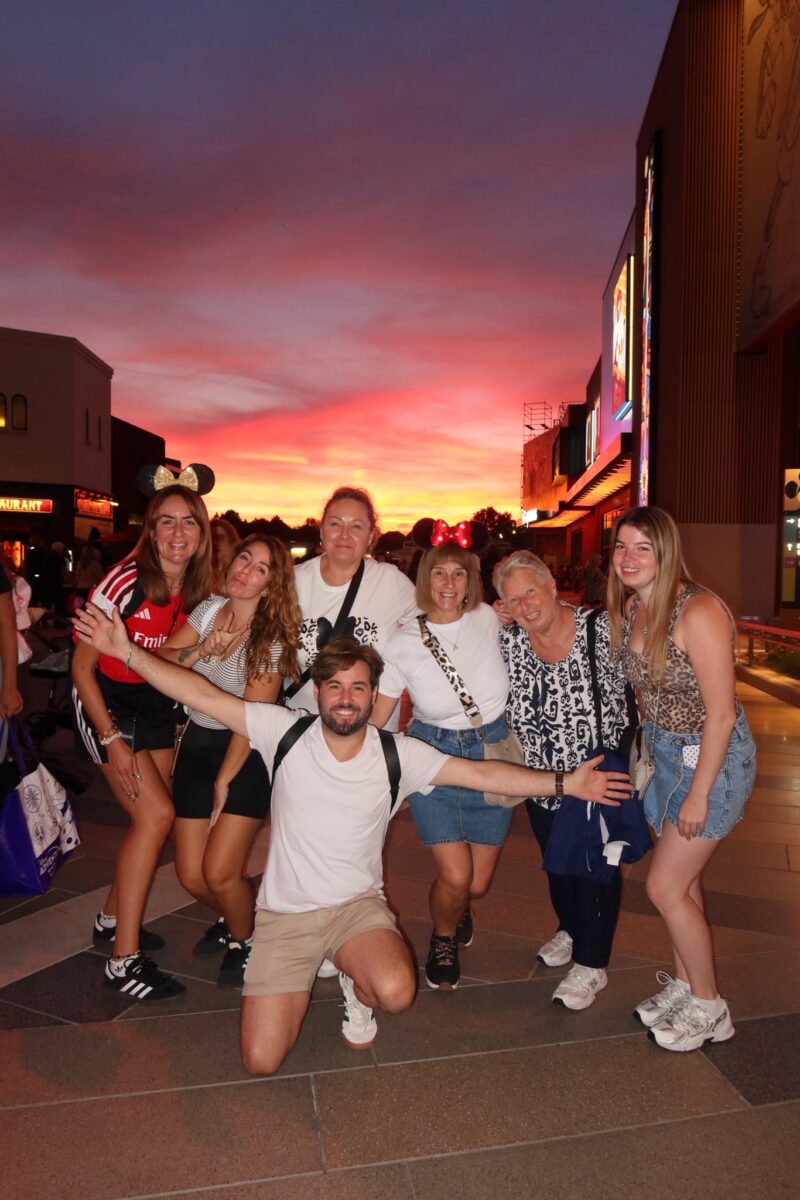 A group of students and staff smile for a photo on a street at sunset, with a pink and orange sky behind them.