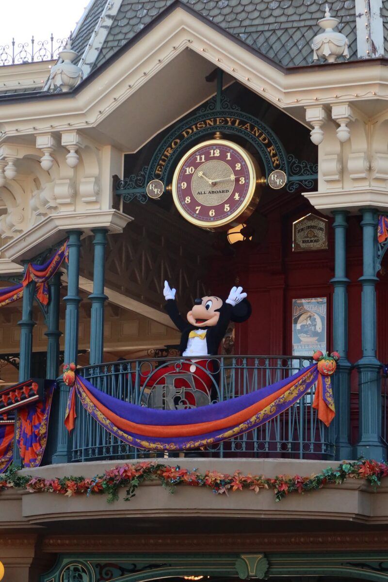 A costumed Mickey Mouse character waves in front of an ornate building with a large decorative clock at a theme park.