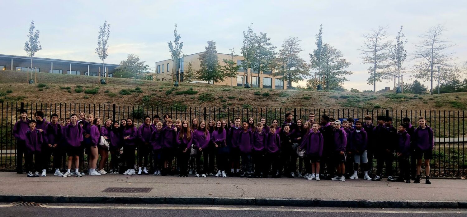 A large group of students in matching purple tops stands in a line on a path, with trees and a building behind them.