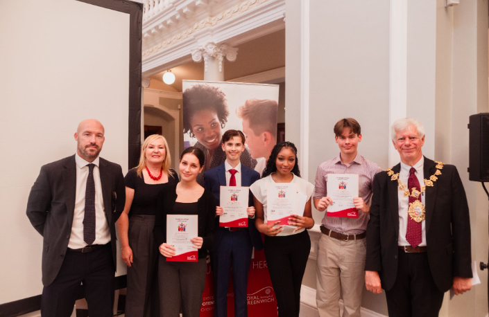 Four young people hold up certificates at an awards event, standing alongside three adults, including a man wearing a mayoral chain of office. They are in a formal room with decorative plasterwork, in front of a Royal Borough of Greenwich banner.