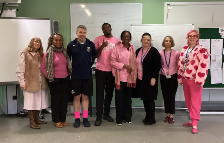 Eight staff members stand in a line in a classroom, each wearing pink clothing or accessories, including pink tops, socks and a heart-patterned jumper. One gives a thumbs up. Whiteboards are on the wall behind them.