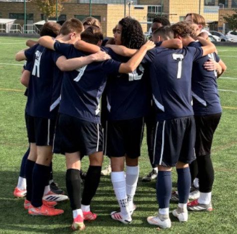 A boys' football team in navy kits stands in a huddle on an artificial pitch, arms around each other's shoulders, with a building behind them.