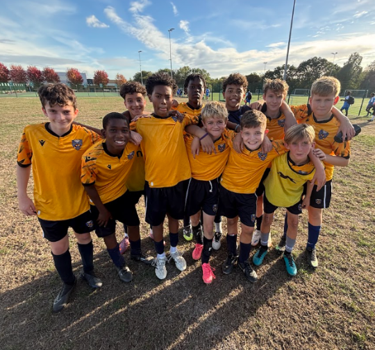 A boys' football team in yellow and black kits poses together on a grass pitch on a sunny day, smiling with arms around each other's shoulders. Trees with autumn leaves and floodlights are in the background.